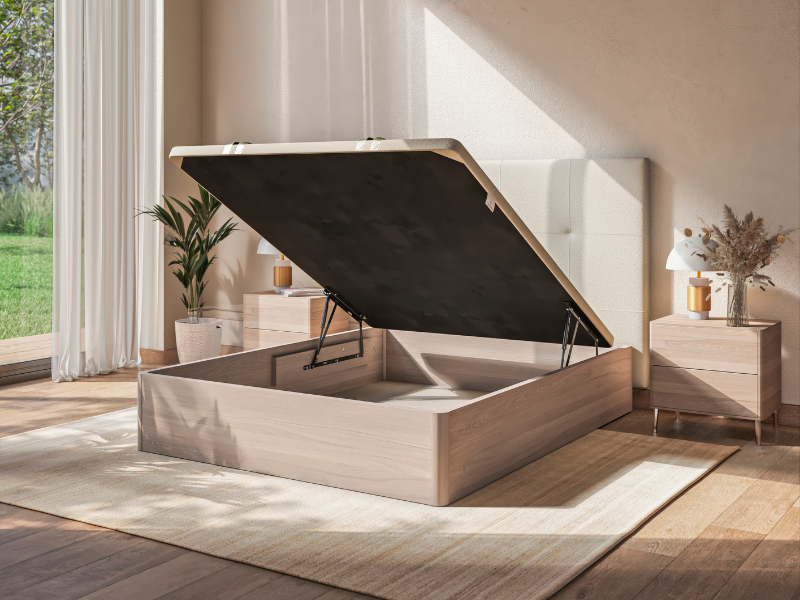 Escena de dormitorio moderno con un canapé abatible levantado mostrando el espacio de almacenamiento, con estructura de madera, cabecero tapizado en gris y mobiliario minimalista